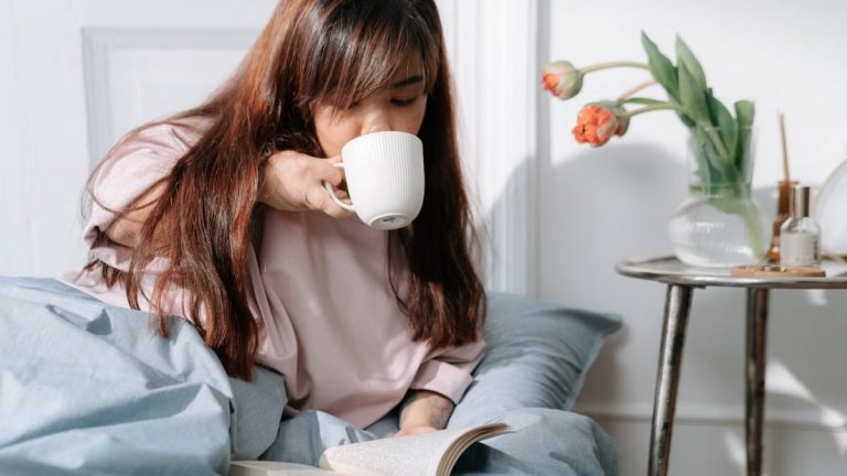 An Asian woman enjoying her morning with coffee and a book in a cozy bedroom setting.